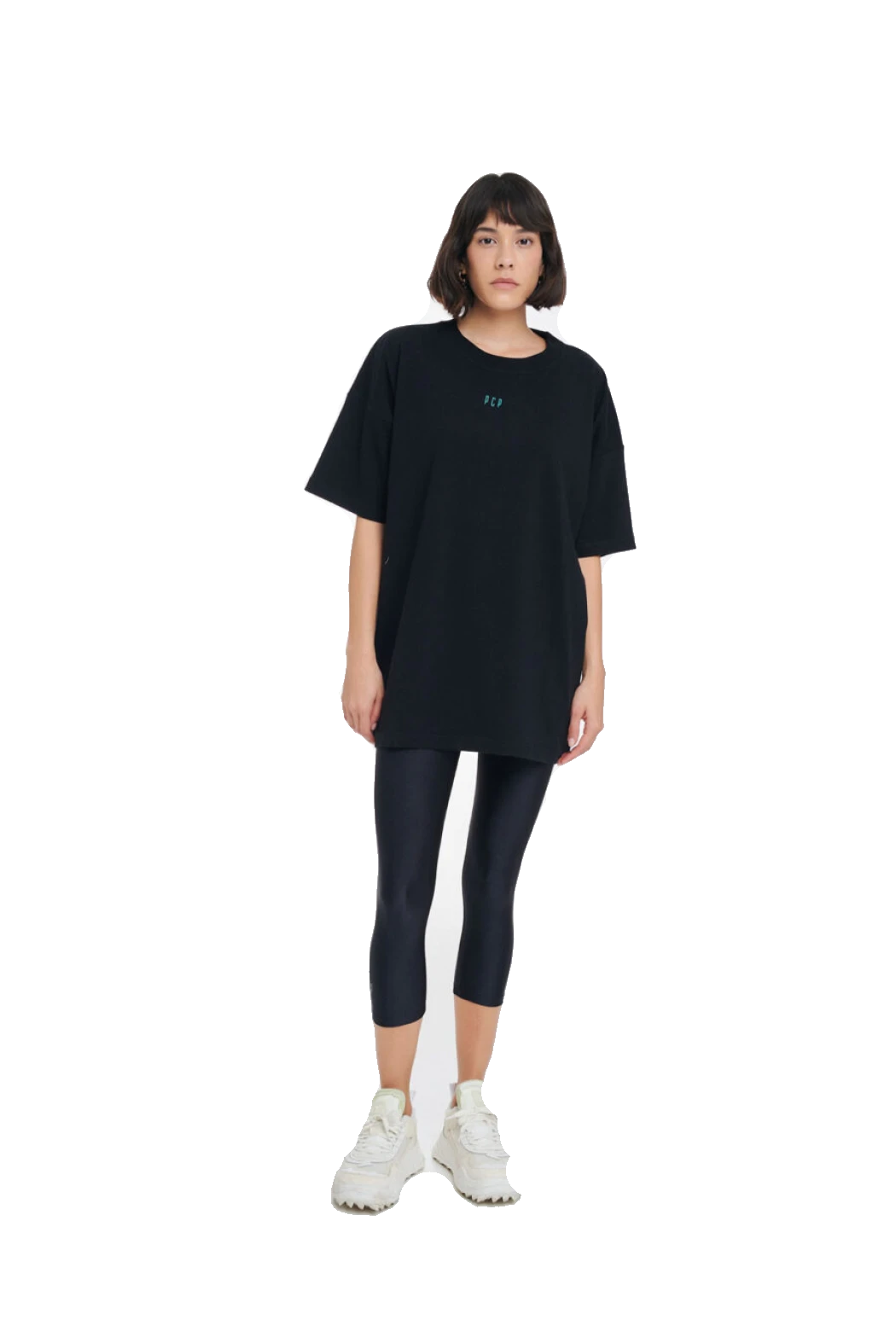 A model stands against a solid white background wearing an oversized black t-shirt with a small blue logo on the chest, black capri leggings, and white sneakers.