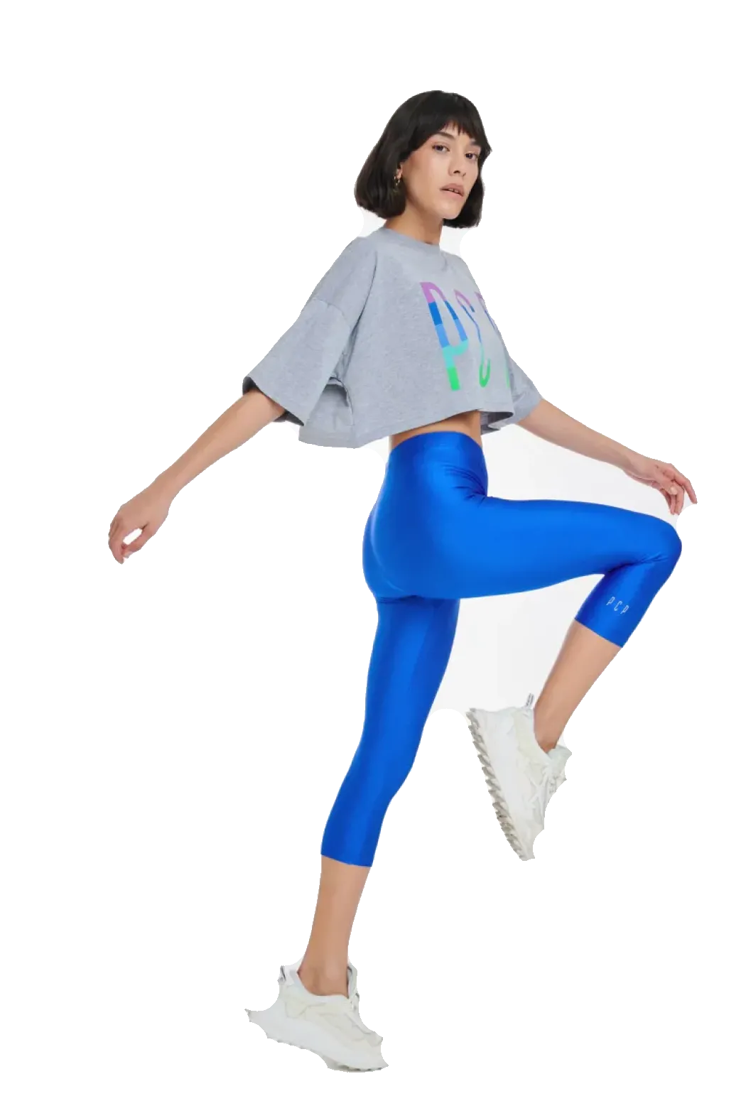 A woman posing in a studio setting, wearing a light gray cropped t-shirt with a colorful graphic, bright blue high-waisted capri leggings, and white sneakers against a solid white background.