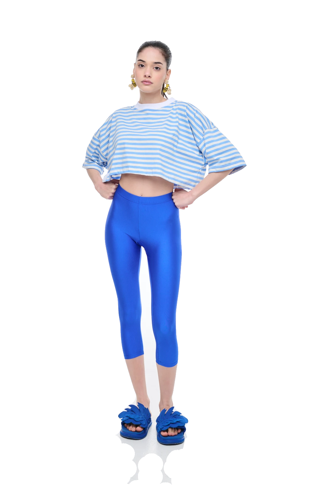A model stands against a white background wearing a light blue and white striped cropped t-shirt, shiny royal blue capri leggings, and matching blue slide sandals.