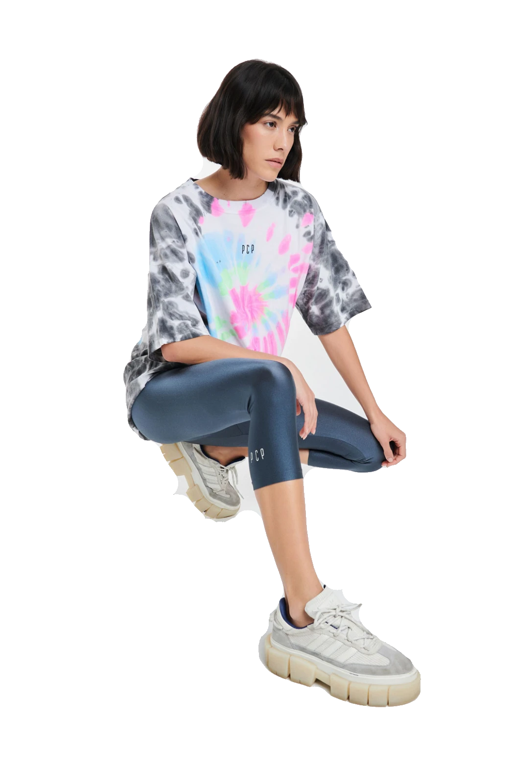 A model wearing a pastel tie-dye t-shirt, dark blue leggings, and chunky white sneakers, crouching in a studio setting against a solid white background.
