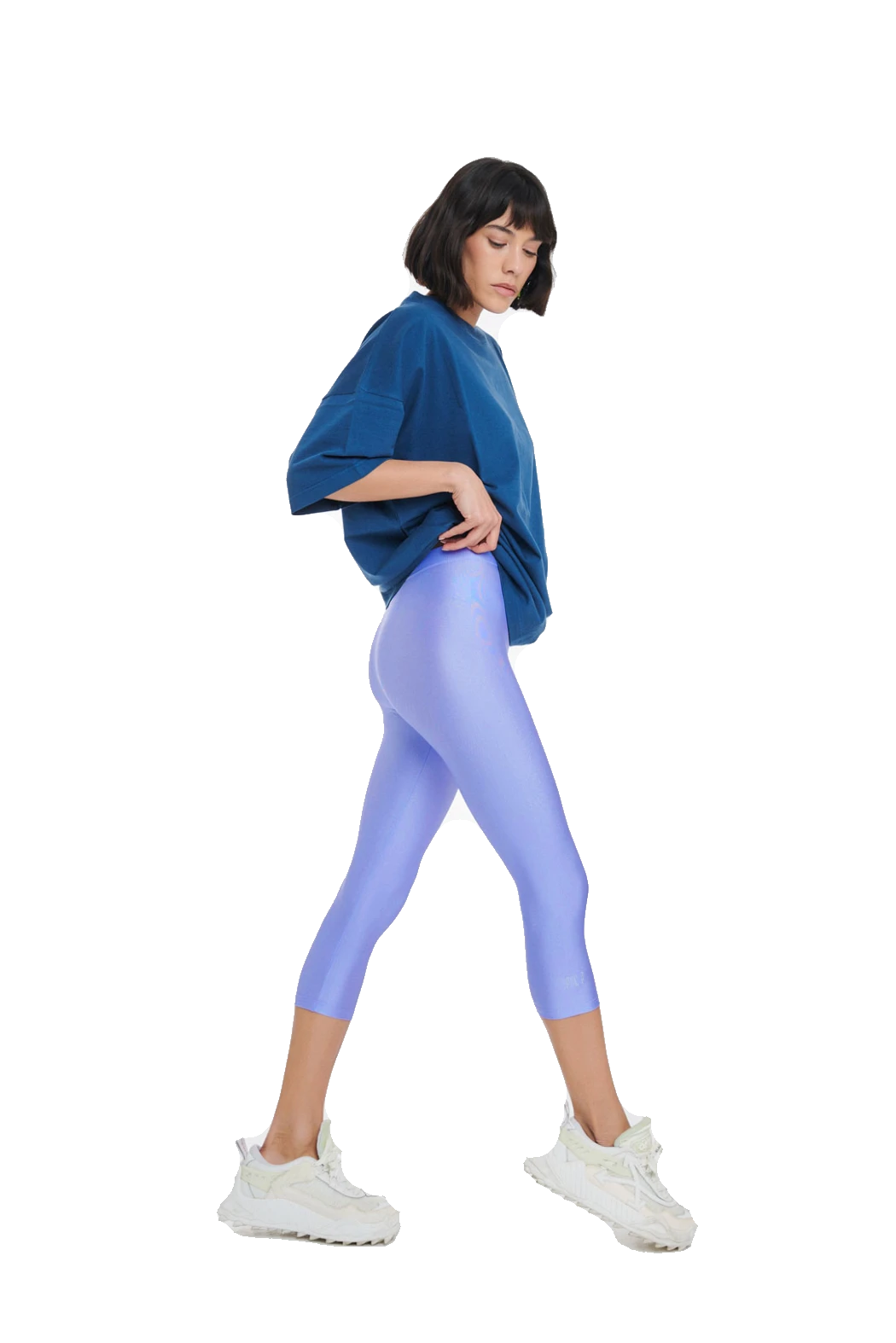 A woman modeling a pair of shiny light purple capri leggings, paired with a blue oversized t-shirt and white sneakers, shown from a side profile against a solid white background.