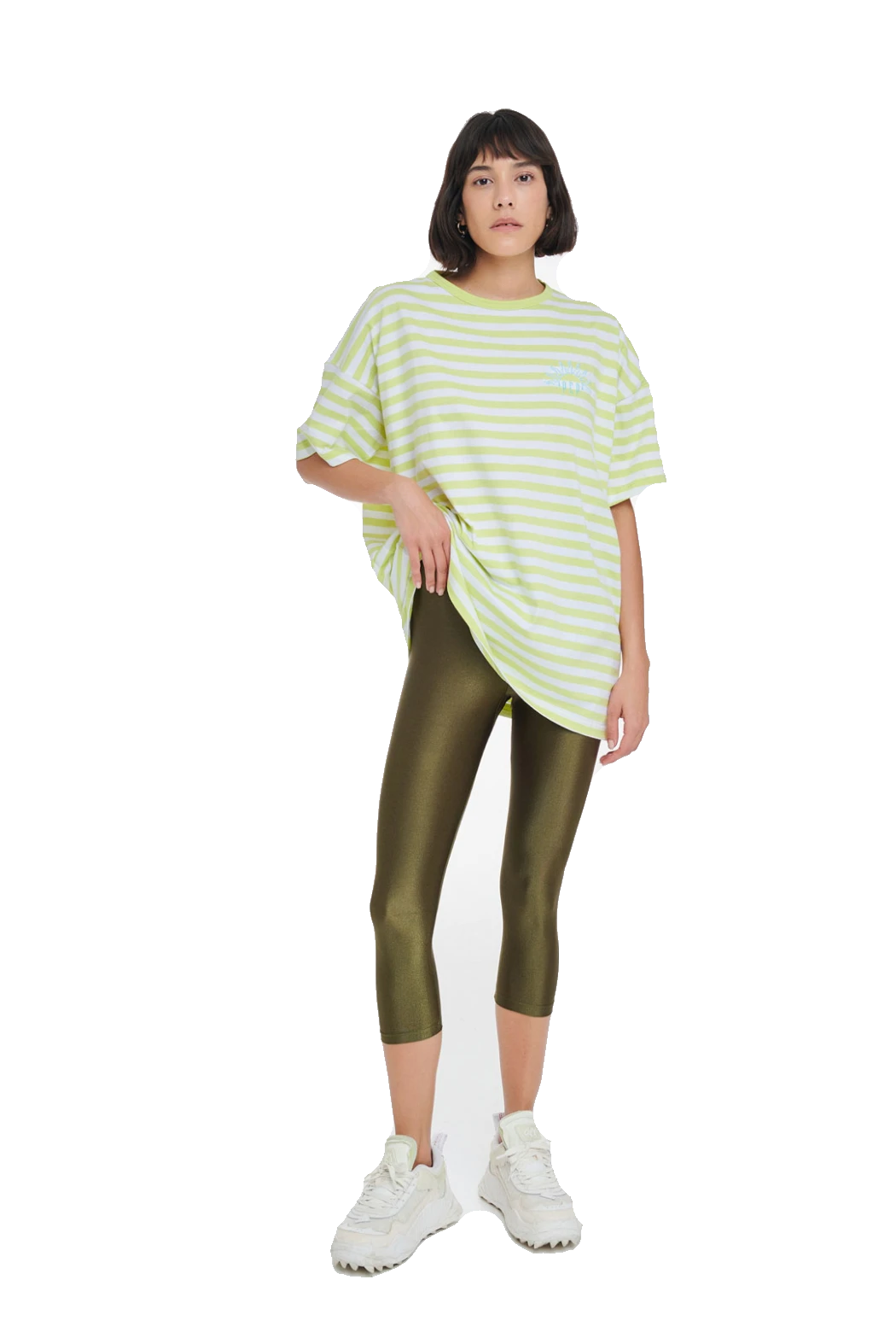A model wearing a light green and white striped oversized t-shirt, shiny olive green capri leggings, and white sneakers, standing against a plain white background.