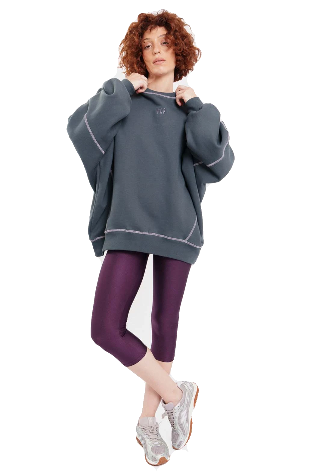 A model with curly red hair wearing an oversized dark slate gray sweatshirt with light gray piping, paired with purple leggings and gray sneakers, against a solid white background.