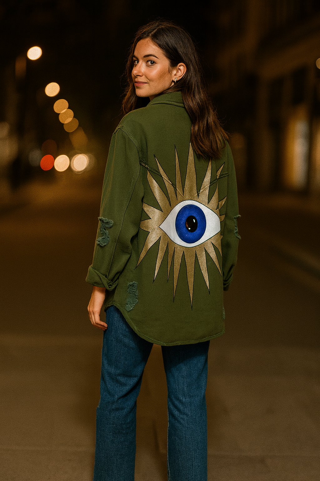 Woman wearing a green jacket with a sun and eye design on the back, standing in a blurred city street at night.