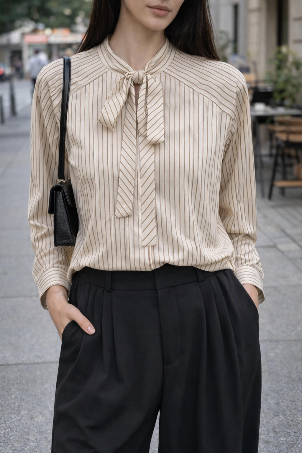 Woman wearing a beige ribbed blouse with a tie detail and black pants on a city street.