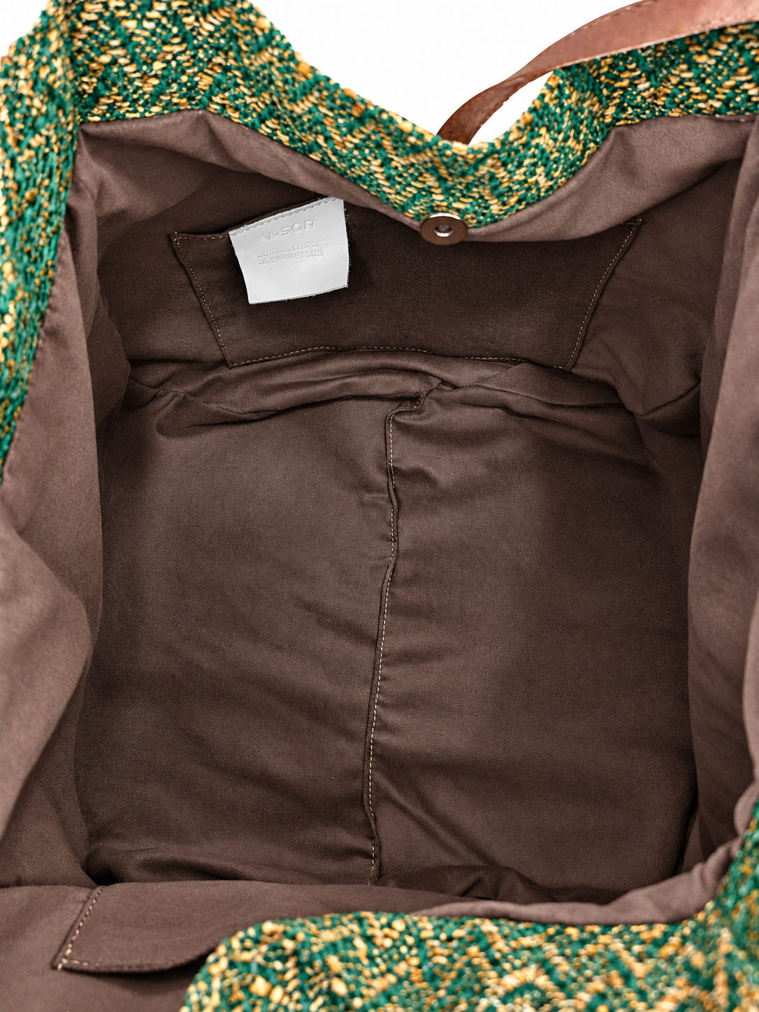 Close-up of the interior of a handbag, showing a brown fabric lining, a small white interior label, and the edge of a green and gold patterned woven material at the top and bottom.