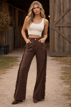 Woman wearing a white crop top and brown fringed pants standing in front of a wooden building.