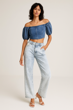 Woman wearing a denim off-shoulder top and light blue jeans on a plain background