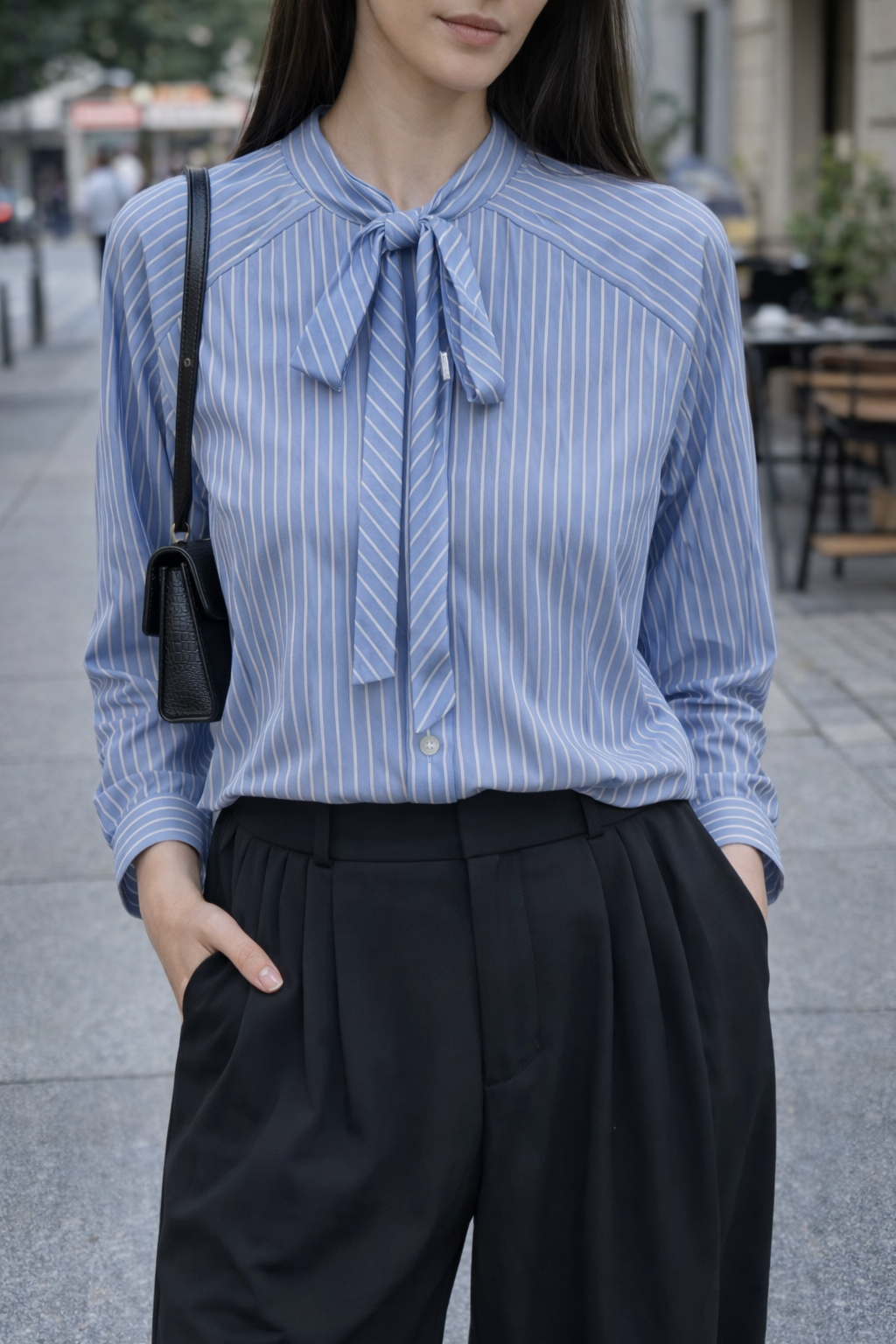 Person wearing a blue striped shirt with a tie and black pants on a city street.