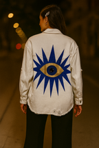 Person wearing a white "Eye" See You jacket with a blue and gold eye design on the back, standing in a blurred outdoor setting.