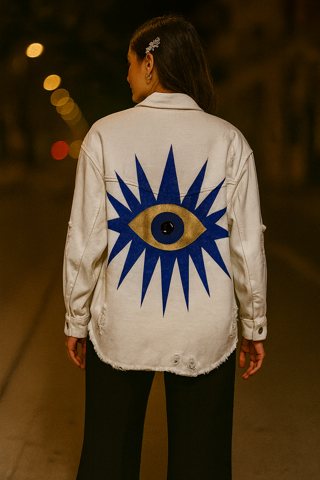 Person wearing a white jacket with a blue and gold eye design on the back, standing in a blurred outdoor setting.