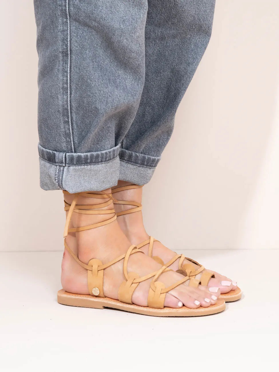 Tan strappy sandals worn with rolled-up jeans on a neutral background