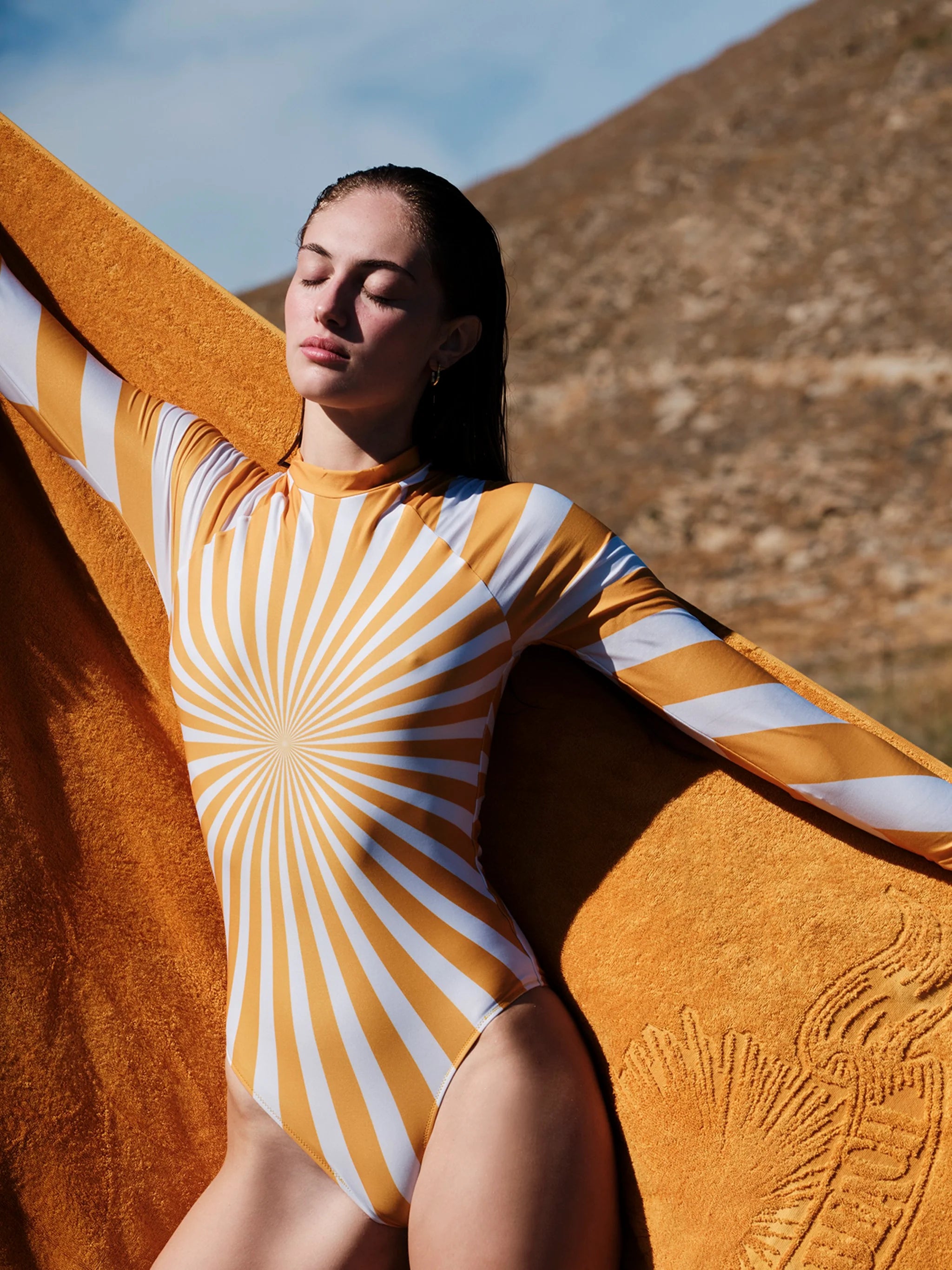 A woman outdoors wearing a fitted long-sleeve bodysuit with a yellow and white sunburst stripe pattern, holding a large textured yellow towel or blanket behind her.