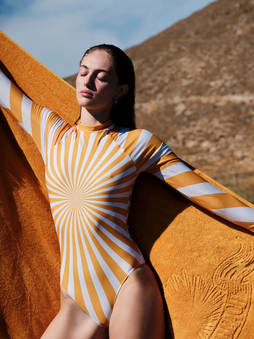 A woman outdoors wearing a fitted long-sleeve bodysuit with a yellow and white sunburst stripe pattern, holding a large textured yellow towel or blanket behind her.