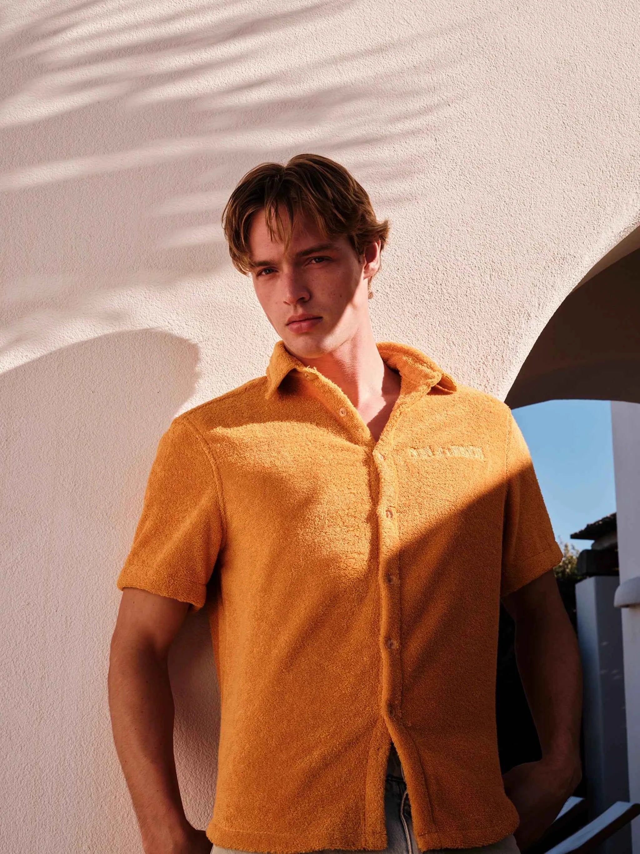 Man wearing a mustard yellow shirt against a textured white wall.