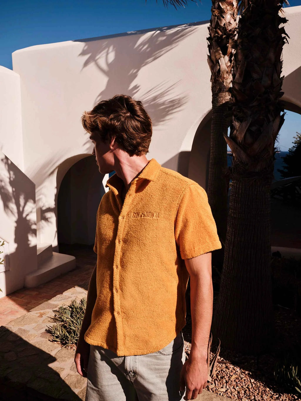 Man wearing a mustard yellow shirt standing in front of a white architectural structure with palm trees.