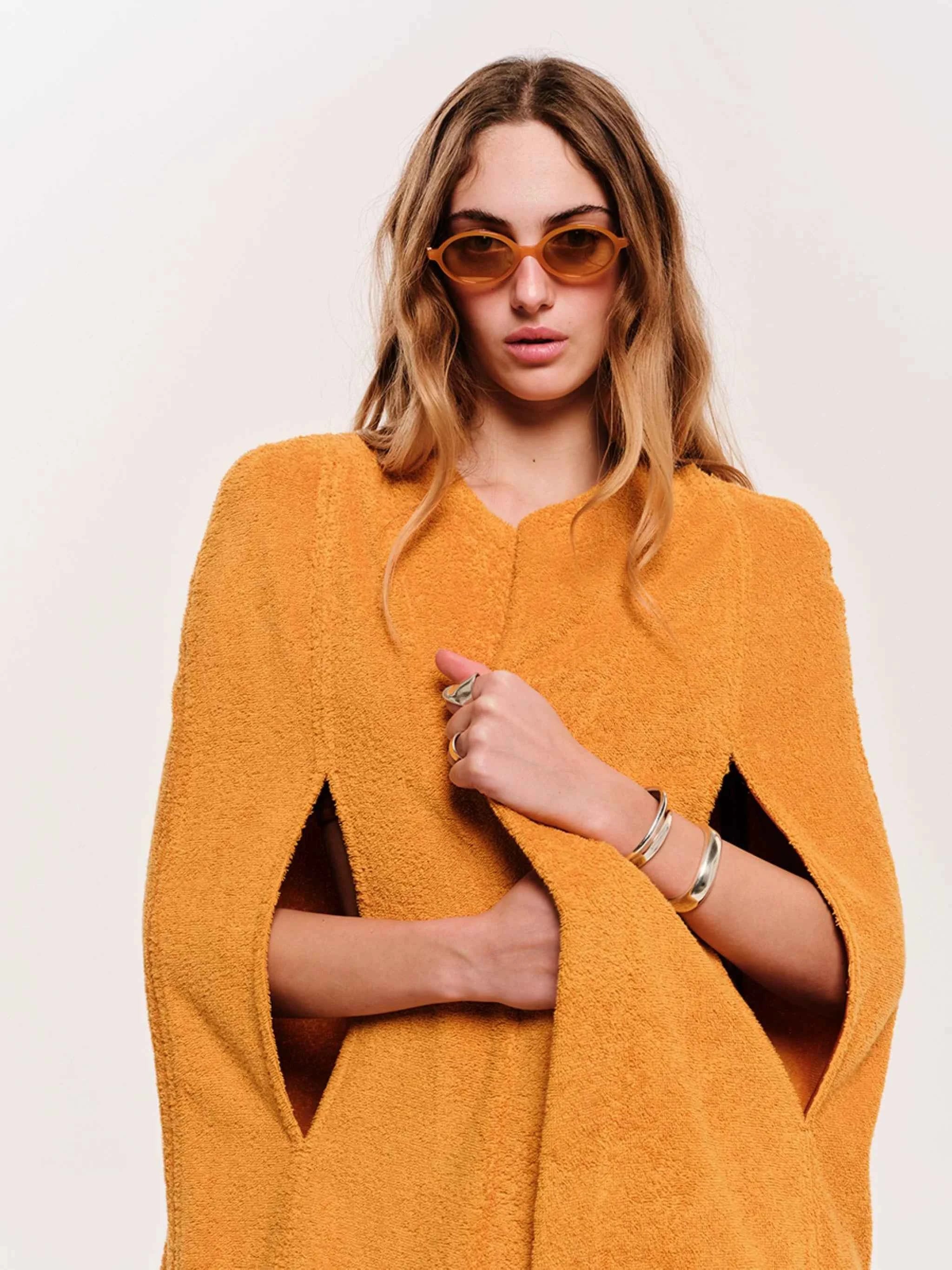 Woman wearing a mustard yellow cape and sunglasses on a plain background