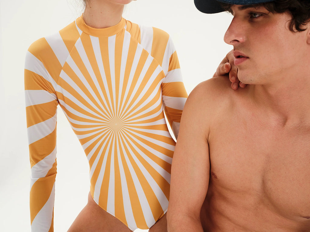 A long-sleeve one-piece swimsuit or bodysuit with a yellow and white sunburst stripe pattern, shown on a model next to a shirtless man against a plain white background.