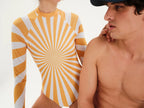 A long-sleeve one-piece swimsuit or bodysuit with a yellow and white sunburst stripe pattern, shown on a model next to a shirtless man against a plain white background.
