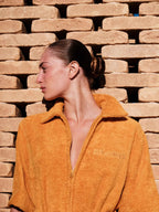 Person wearing a mustard yellow jacket with a brand logo against a brick wall.