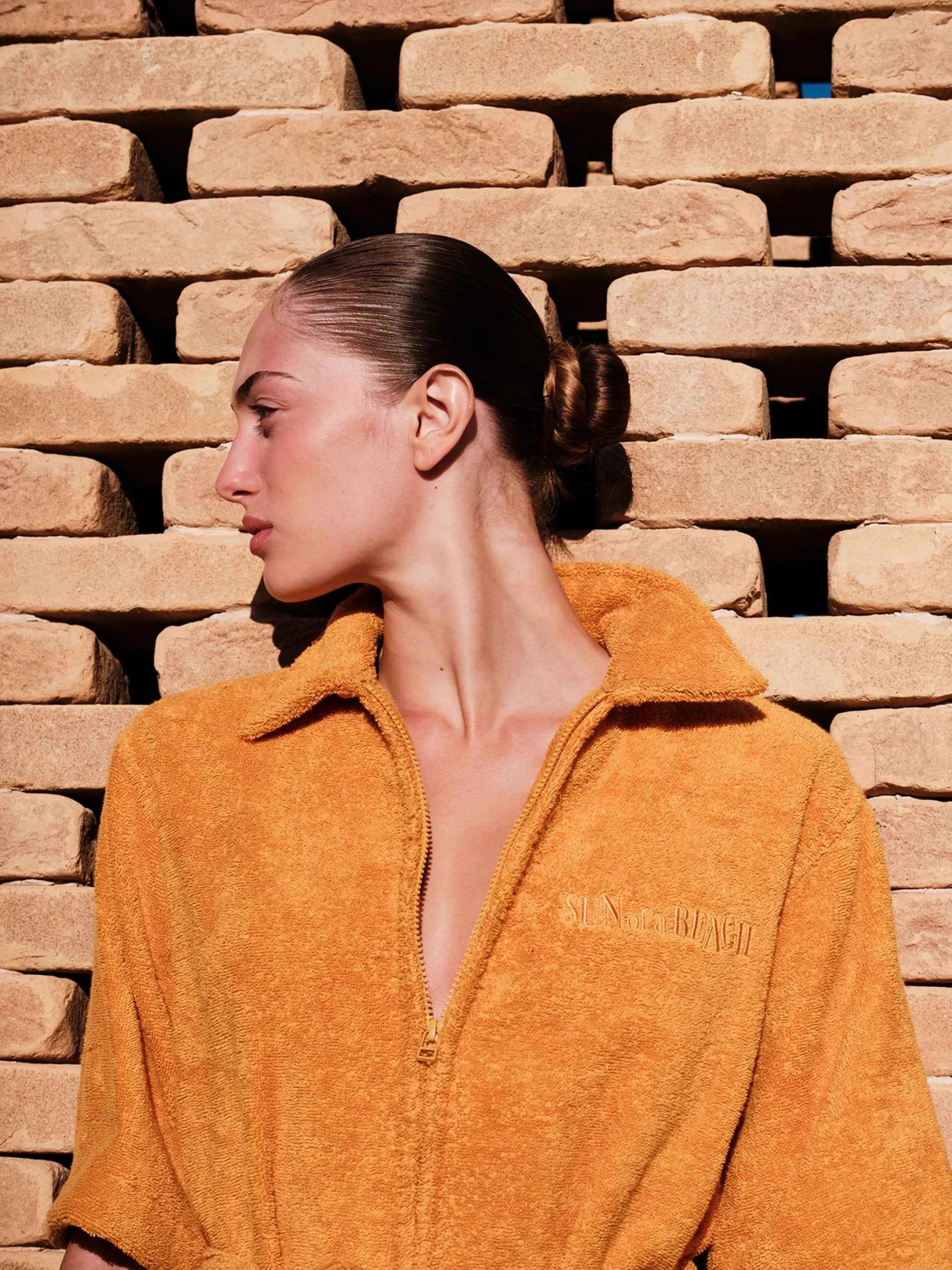 Person wearing a mustard yellow jacket with a brand logo against a brick wall.
