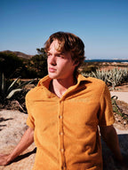 Man wearing a mustard yellow shirt outdoors with plants and blue sky in the background
