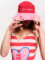 Person wearing a red 'Sun of a Beach' bucket hat and a red and white striped tank top with a heart design.