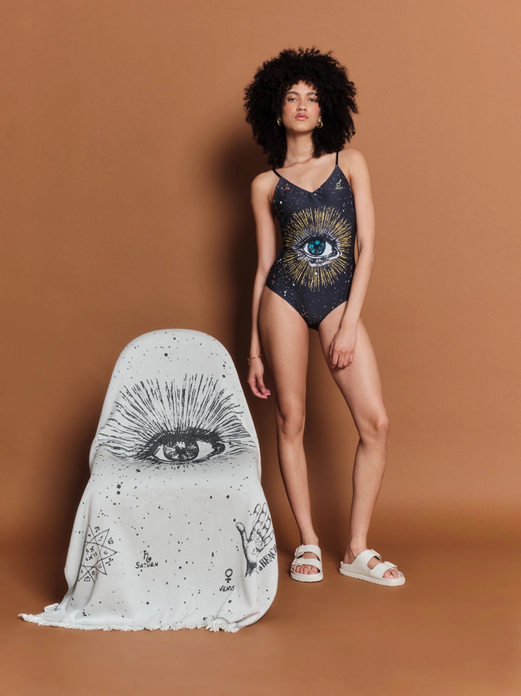 Woman in a swimsuit with eye design standing next to a chair with similar design against a brown background