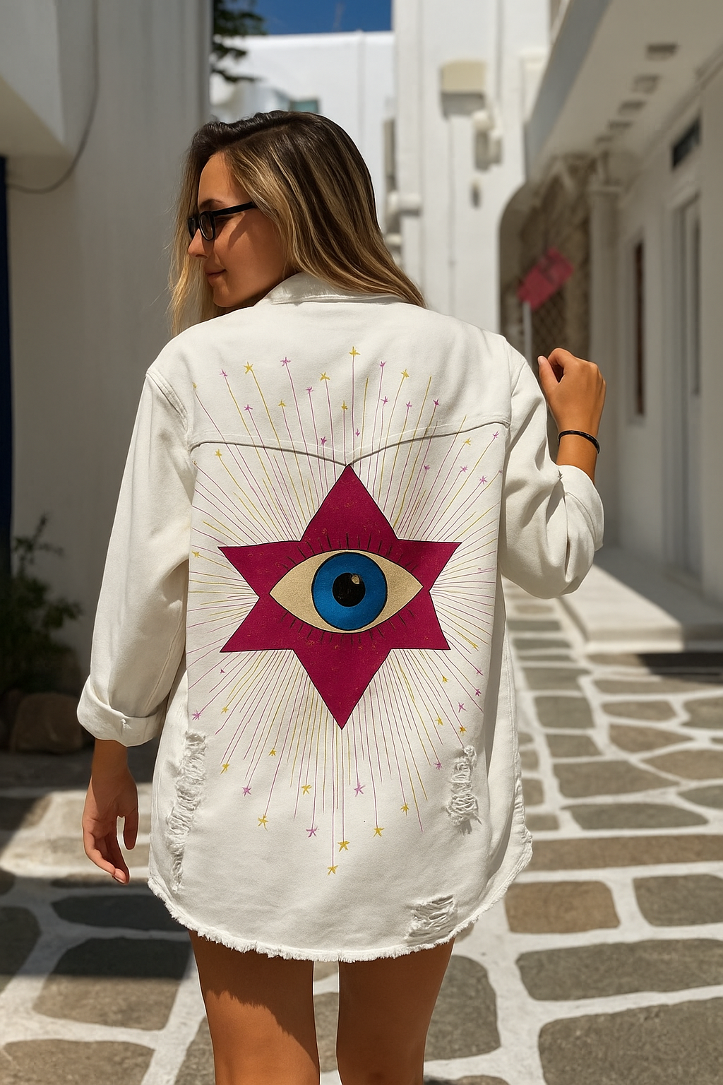Woman wearing a white denim jacket with a star and eye design in a narrow street.