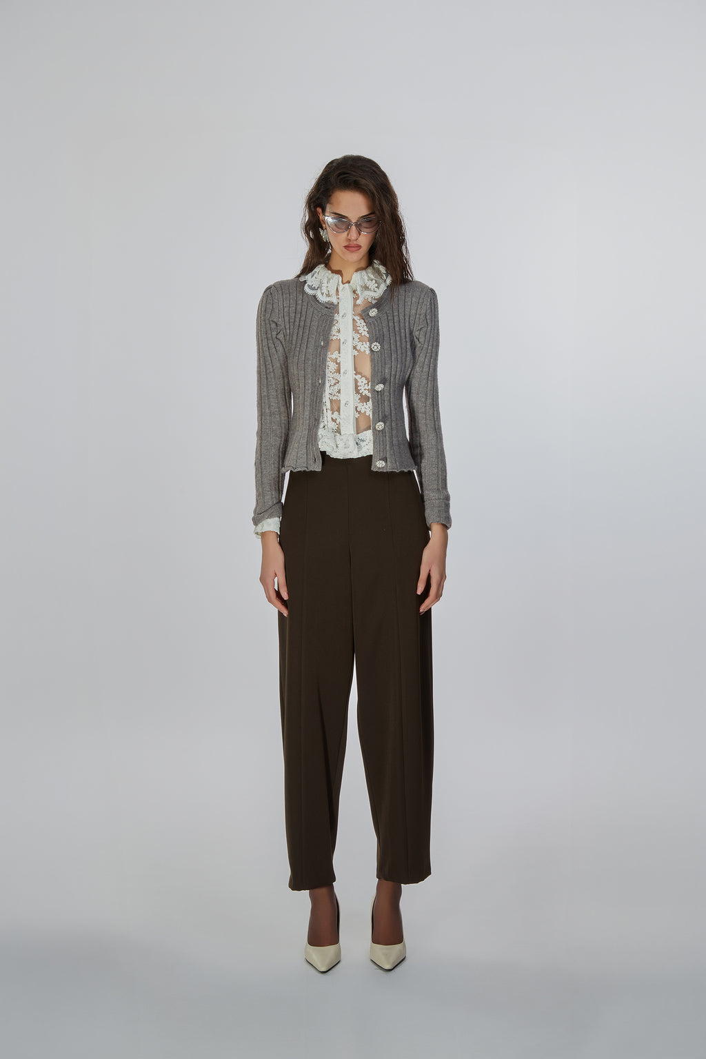 A model stands against a light gray studio background wearing a gray ribbed knit cardigan over a white ruffled blouse with a floral print, paired with dark brown cropped trousers and beige pointed-toe heels.
