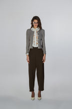 A model stands against a light gray studio background wearing a gray ribbed knit cardigan over a white ruffled blouse with a floral print, paired with dark brown cropped trousers and beige pointed-toe heels.