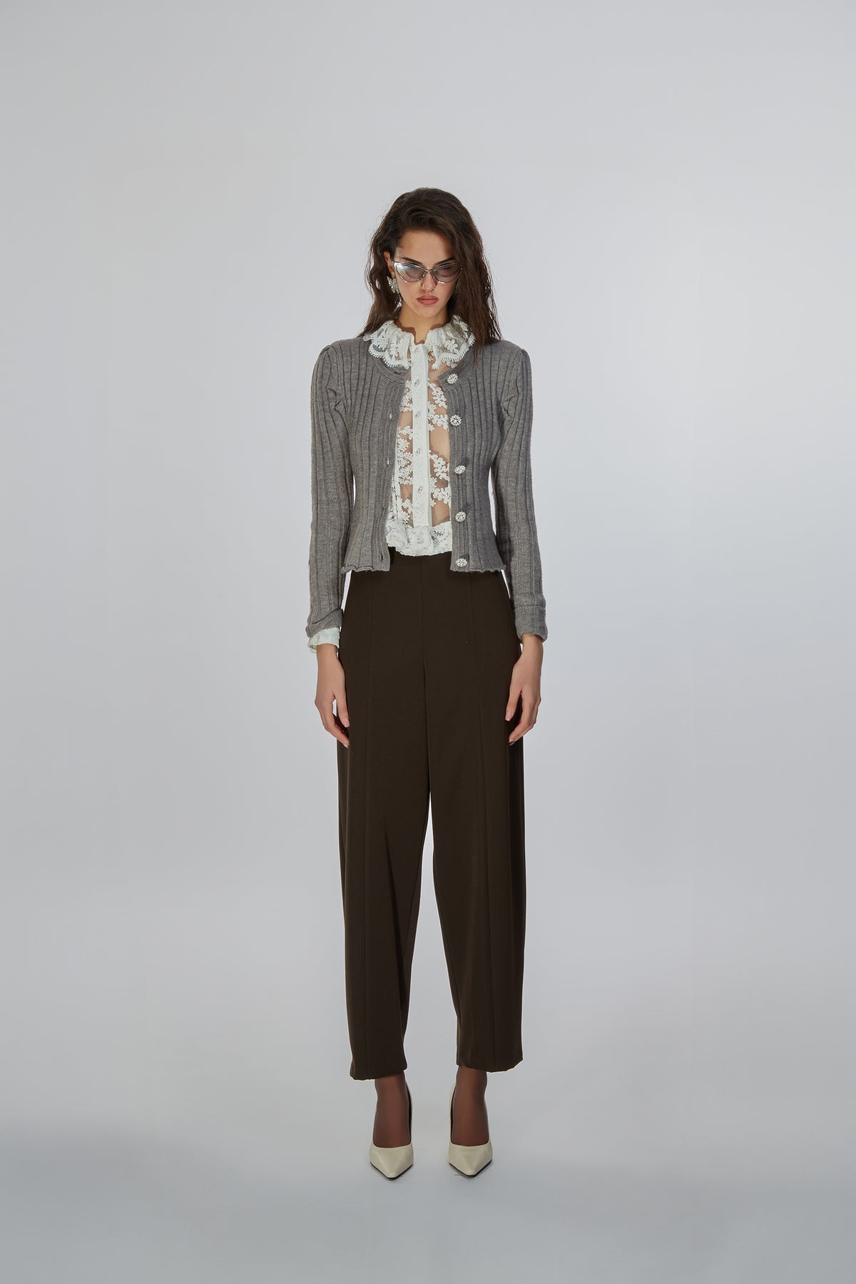 A model stands against a light gray studio background wearing a gray ribbed knit cardigan over a white ruffled blouse with a floral print, paired with dark brown cropped trousers and beige pointed-toe heels.