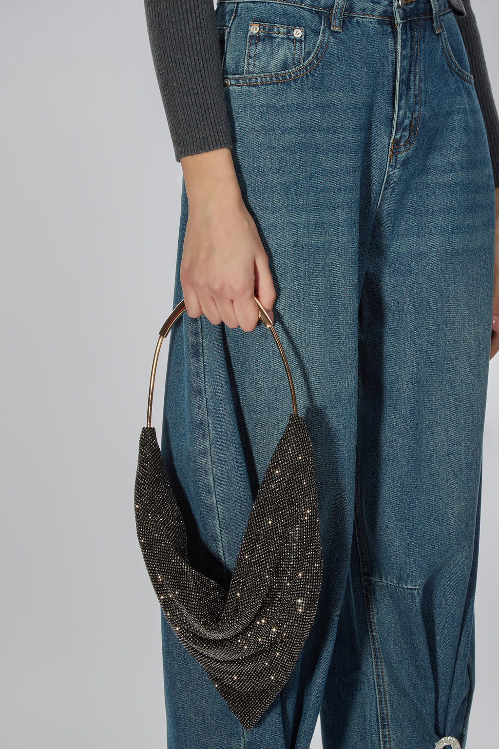 A person wearing blue jeans and a dark gray ribbed long-sleeve top holds a draped black beaded crescent shoulder bag with gold-tone metal handles against a light gray studio background.