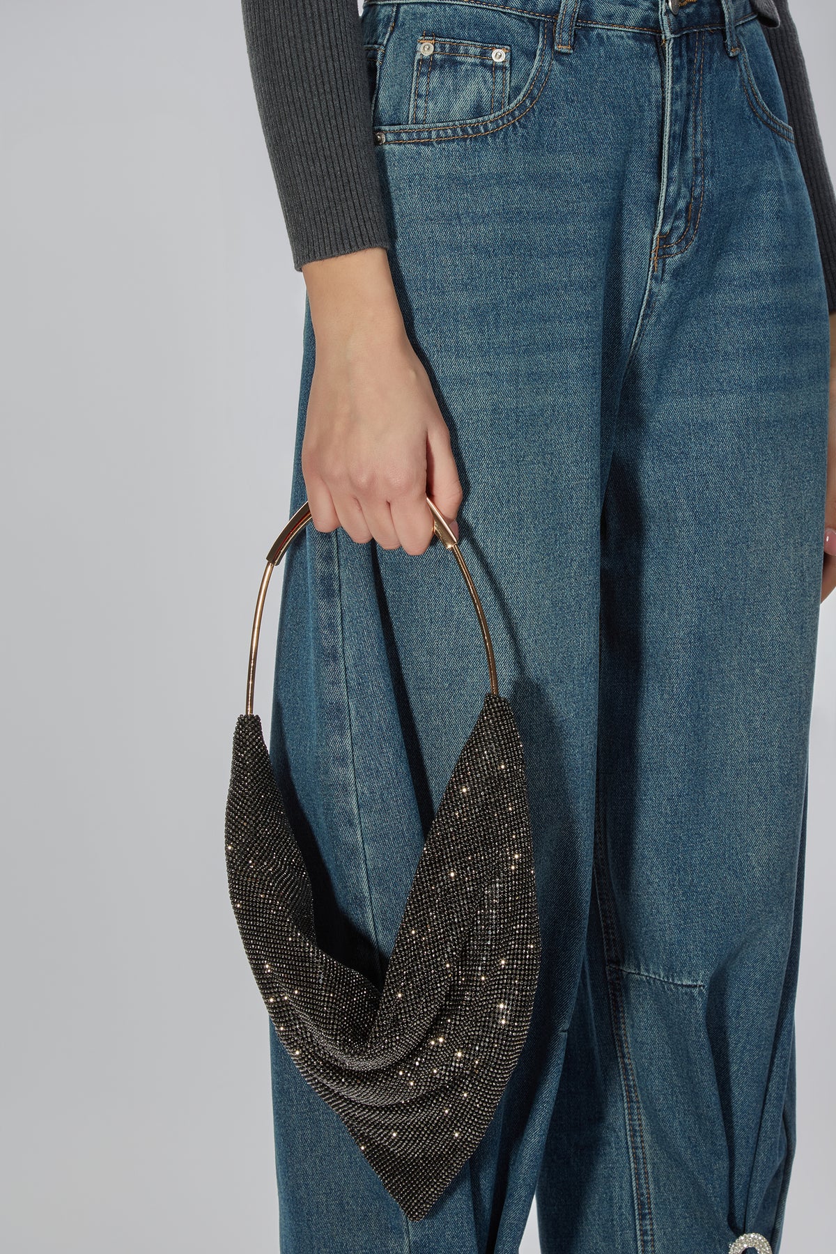A person wearing blue jeans and a dark gray ribbed long-sleeve top holds a draped black beaded crescent shoulder bag with gold-tone metal handles against a light gray studio background.