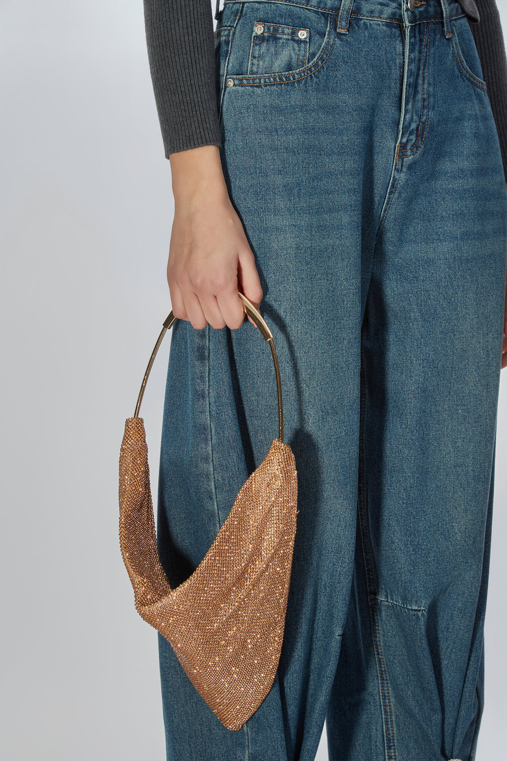 A person wearing blue jeans and a dark gray ribbed top holds a metallic gold glitter crescent-shaped shoulder bag with a gold-tone handle against a light gray studio background.