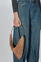 A person wearing blue jeans and a dark gray ribbed top holds a metallic gold glitter crescent-shaped shoulder bag with a gold-tone handle against a light gray studio background.