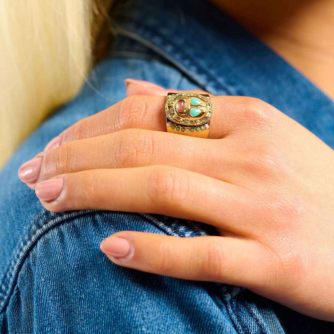 Close-up of a hand wearing a gold ring with gemstones, resting on a denim surface.