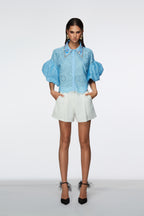 Woman wearing a light blue blouse with ruffled sleeves and white shorts on a white background