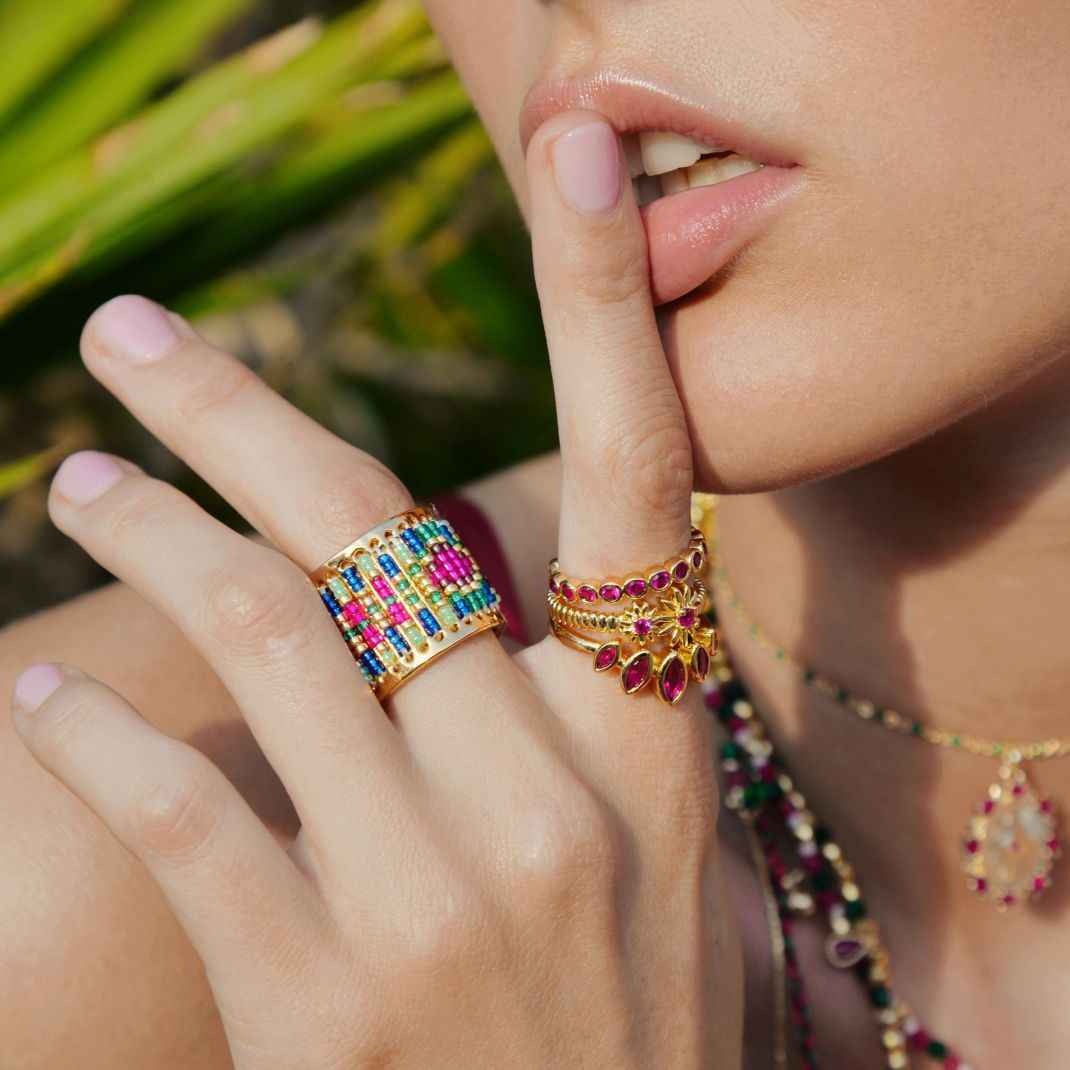 Close-up of a hand with colorful gemstone rings and a necklace, with a blurred natural background.