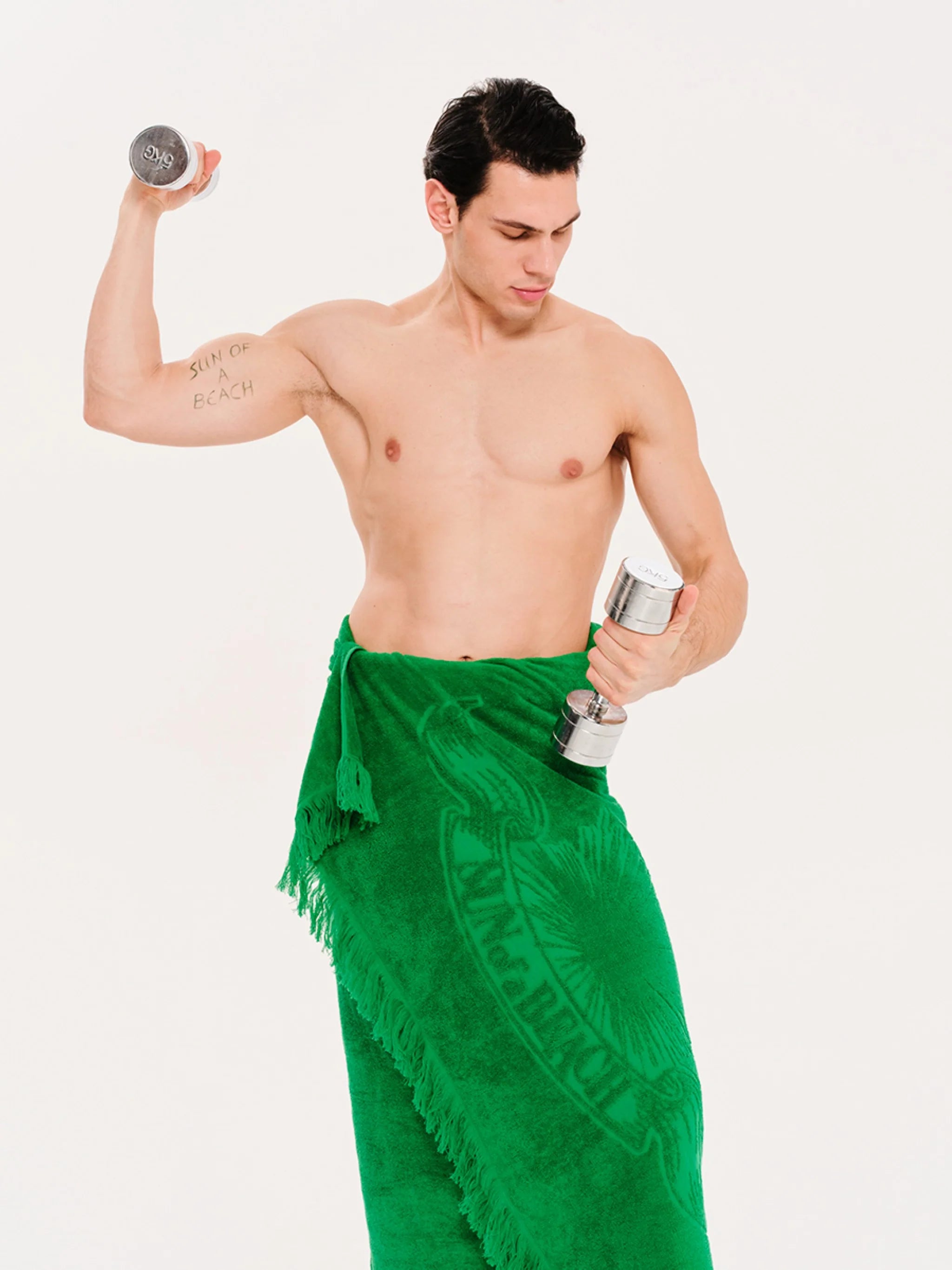 Man holding dumbbells wearing a green towel around his waist on a white background