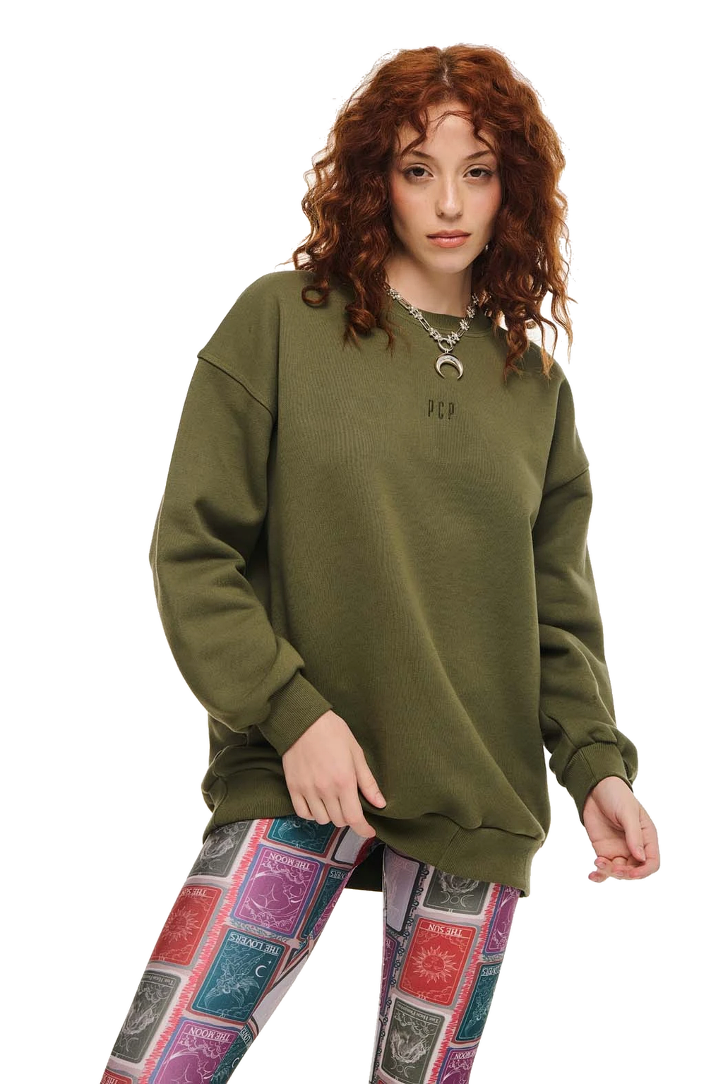 A woman with curly red hair wearing an oversized olive green crewneck sweatshirt and colorful patterned leggings, standing against a plain white background.