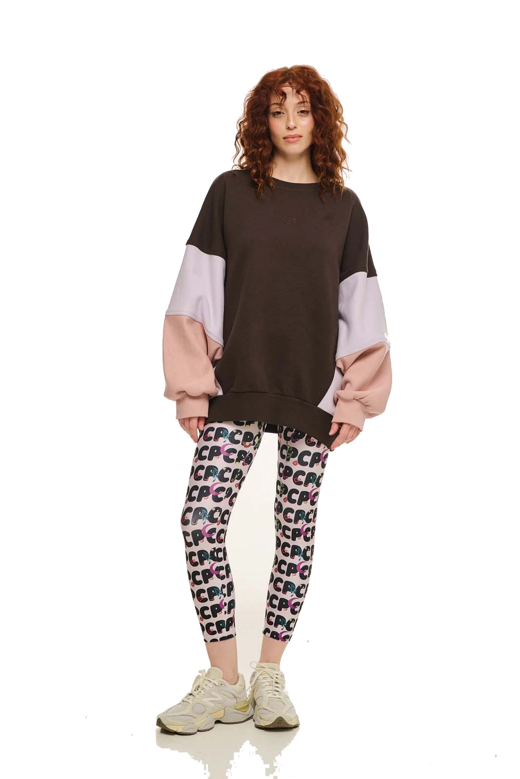 A model with curly red hair stands against a white studio background wearing a color-blocked sweatshirt with a dark brown body, light purple sleeves, and pink balloon sleeves, paired with patterned leggings and beige sneakers.