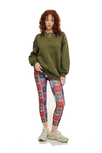 A model with curly red hair stands against a white background wearing an oversized olive green sweatshirt, colorful patterned leggings, and beige sneakers.