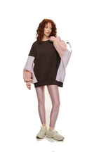 A model with curly red hair stands against a white background wearing an oversized color-block sweatshirt in dark brown, light pink, and pale lavender, paired with shiny pink leggings and beige sneakers.