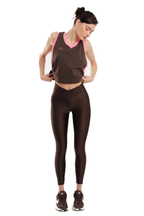 A woman modeling a dark brown athletic set consisting of a cropped tank top with pink trim and matching high-waisted leggings, standing against a plain white background.
