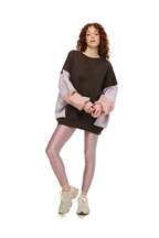 A model wearing a color-blocked oversized sweatshirt with dark brown, light pink, and lavender sleeves, paired with metallic pink leggings and beige sneakers, on a solid white background.