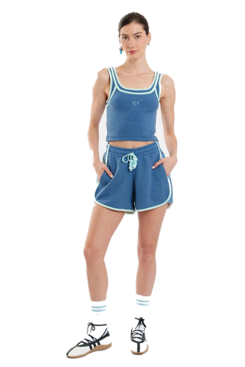 A model wearing a matching blue athletic set consisting of a cropped tank top and shorts with white trim, paired with white socks and black-and-white sneakers, against a plain white background.