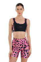 A woman modeling a matching activewear set consisting of a black sports bra with pink side panels and a pink biker shorts set with a black leopard print, on a solid white background.