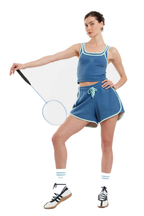 A woman modeling a blue athletic crop top and shorts set with light blue trim, white socks, and black-and-white sneakers, holding a badminton racket against a plain white background.