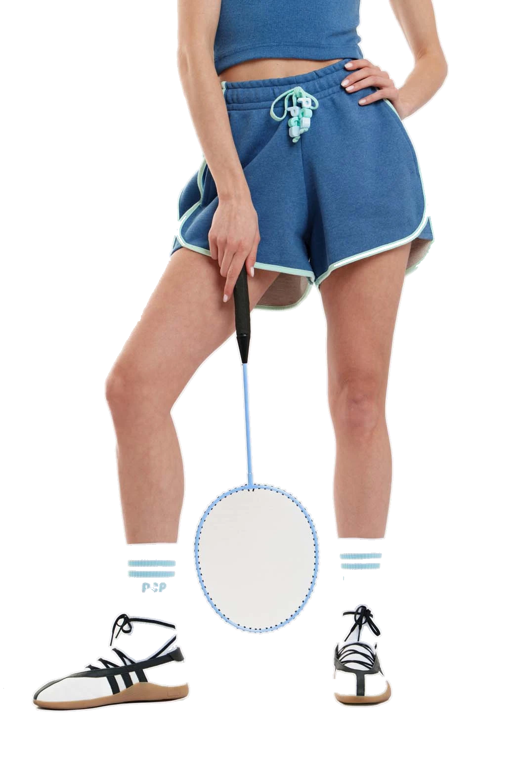 A model wearing blue athletic shorts with white trim and a matching blue crop top, holding a badminton racket. The model is also wearing white socks with blue stripes and black-and-white sneakers.